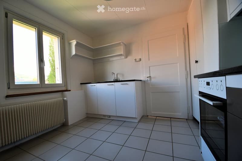 Clean and empty kitchen with white tiles, black and white stove, sink, wooden window and radiator.