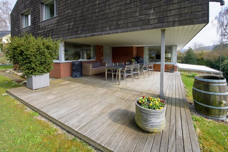 modern house, wooden deck, outdoor dining table, couch, potted plants, barrels