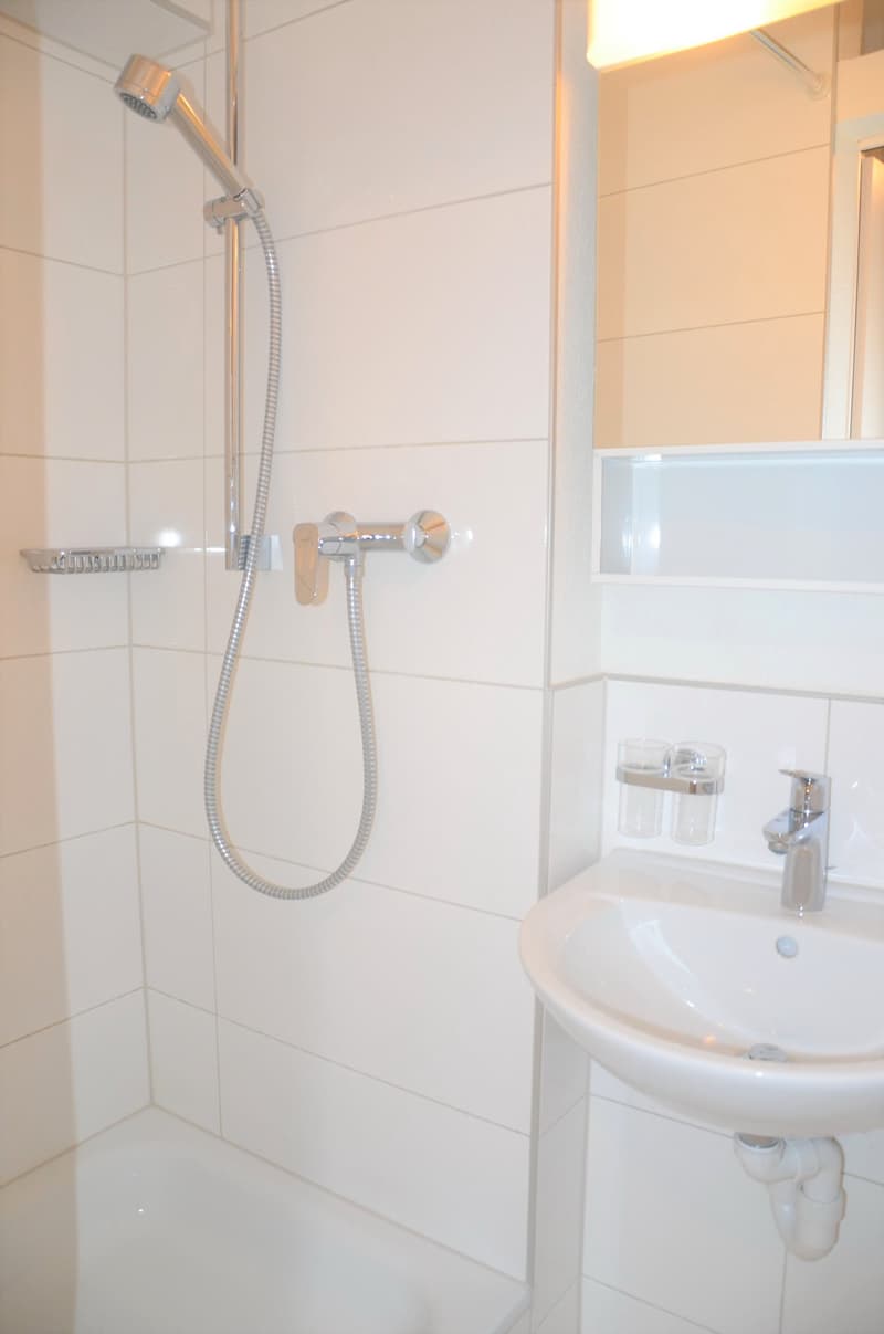 Modern white bathroom with a shower, sink, mirror, and a wall-mounted soap dish.