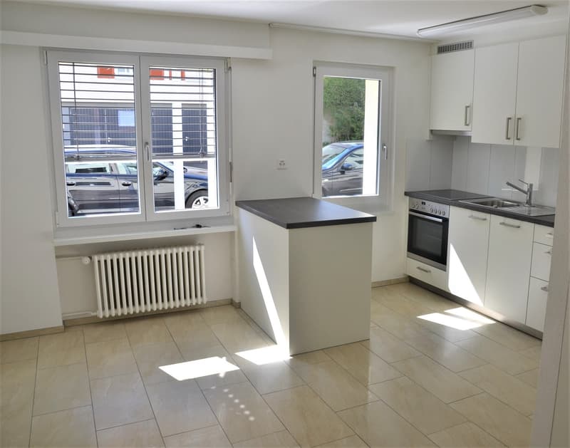 open-plan kitchen with light tiles, white kitchen units, black counter top, large window with view to parking lot, white radiator, white ceiling light