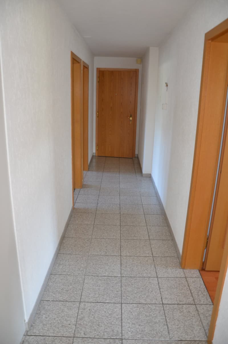 Empty hallway with tiled floor, white walls, wooden doors, and a power outlet on the right wall.