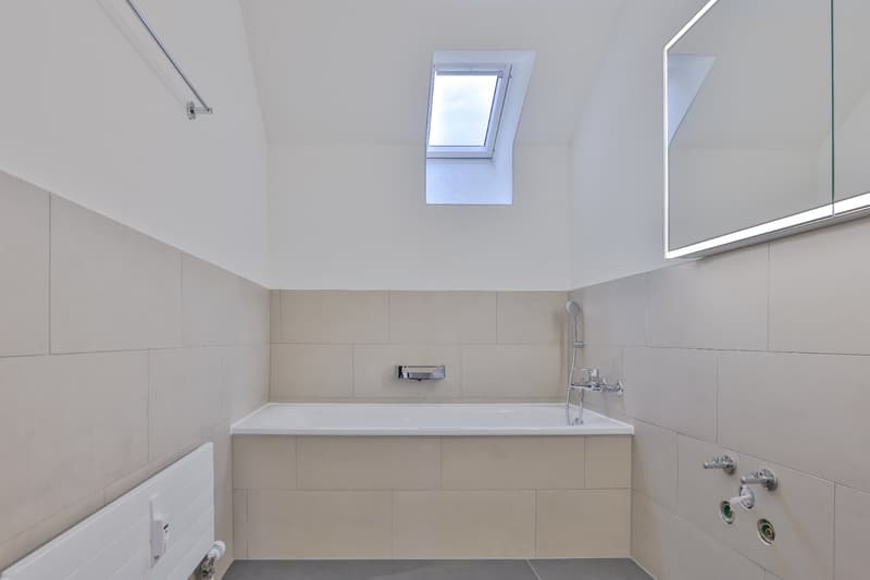A clean and minimalistic bathroom with beige tiles, white bathtub, glass mirror, and a skylight.