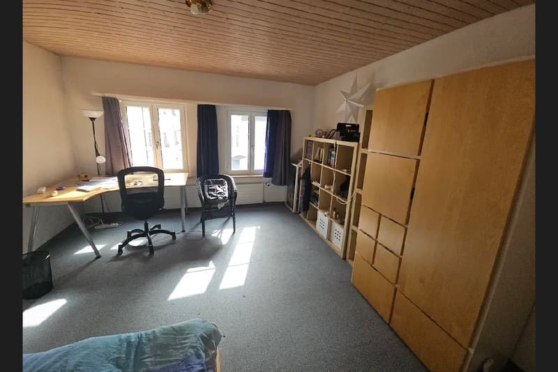 Spacious room with desk, chairs, and shelves. Desk has a lamp, chair, and table, shelves with boxes and books. Bed at the bottom, trash can, and a white star on the wall.