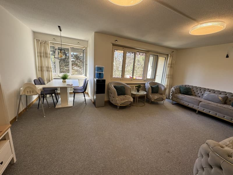Dining room with a table, chairs, and a sofa. There is a potted plant on the table, and a water dispenser is placed on the floor. The room has a carpeted floor, and there are two lamps mounted on the ceiling. The room has a window with curtains, and there