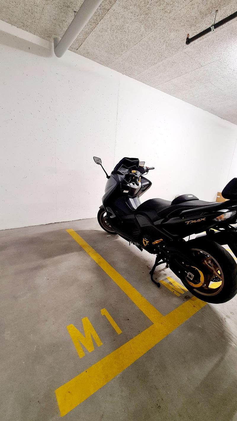 Underground parking space with a motorcycle parked in it, yellow parking lines, white walls, grey floor