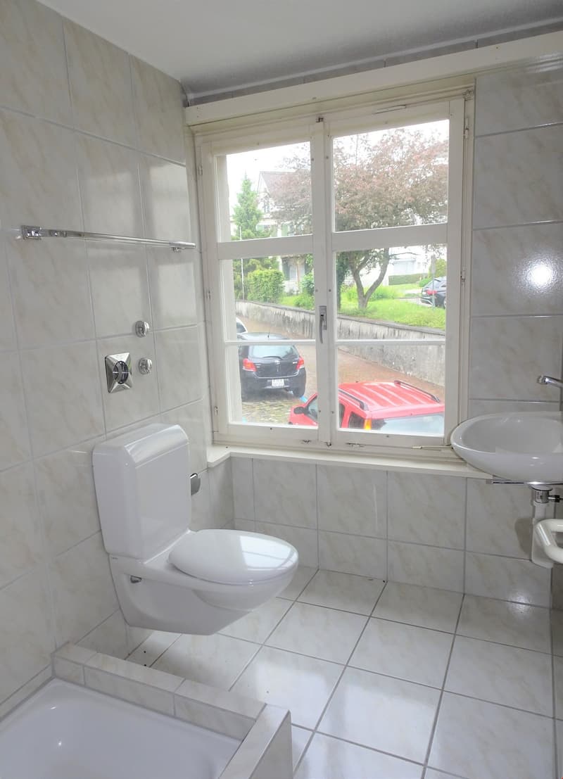 Tiled bathroom with toilet, sink, and large window overlooking a parking area and outdoor scenery