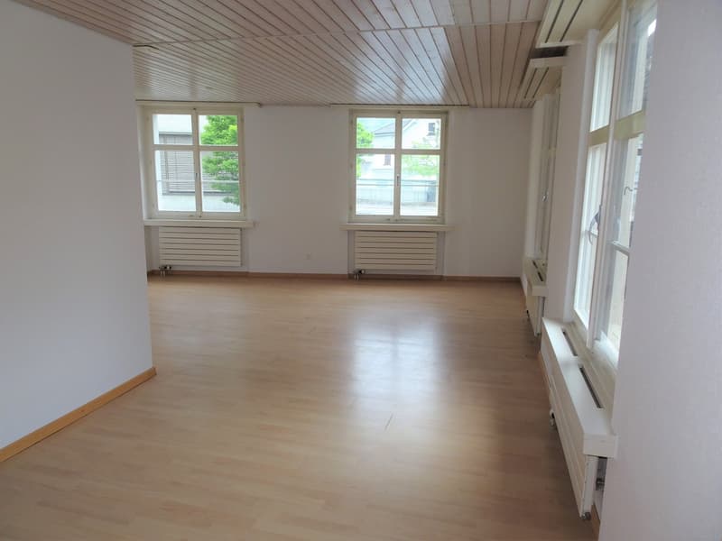 Spacious room with wooden flooring, white walls, and large windows providing natural light. The room has a vaulted ceiling with wooden paneling.