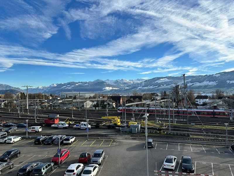 Outdoor parking lot with rows of parked cars and electric poles, railway tracks, red train on tracks, snow-capped mountains in the background.