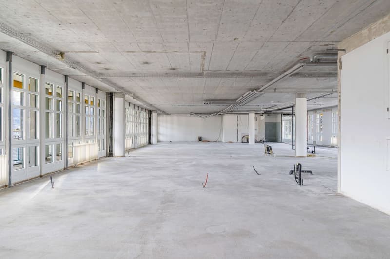 An empty, unfinished room with a large open space, concrete floor, large glass windows, white walls, and exposed utility pipes.