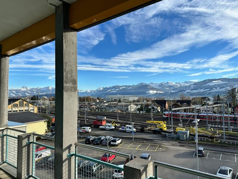 balcony with metal railing, clear view of mountains, parking lot with many cars, railway with trains and maintenance vehicles
