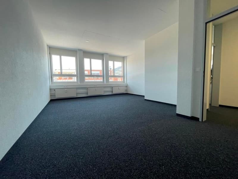 Large open office space with white walls, multiple windows, and dark carpeted flooring.