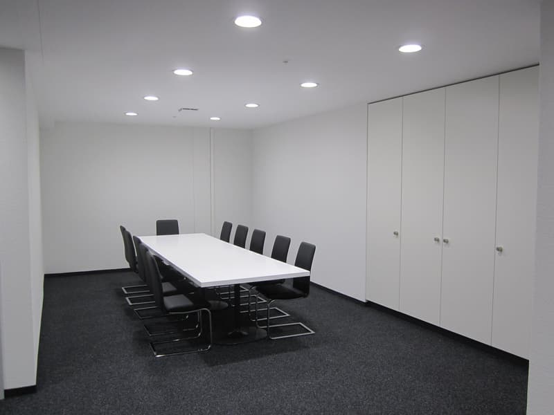 A large, minimalist conference room with a long white table, black chairs, and built-in storage cabinets along the walls. The room is well-lit with recessed ceiling lights.