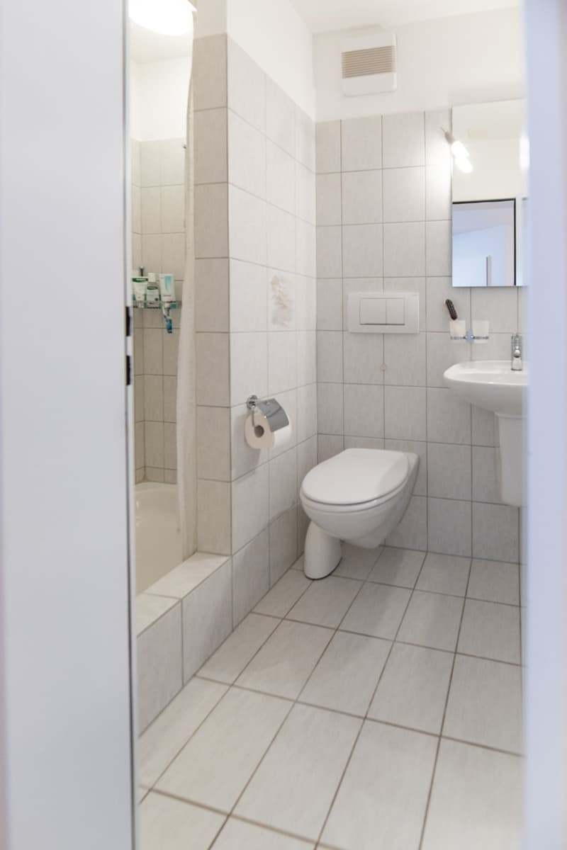 Bathroom with white tiles, toilet, sink, shower, mirror, and a small shelf