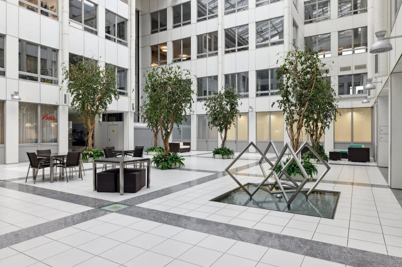 Large open atrium, white tile floors, white walls, decorative trees, seating area with tables, water feature, mounted lighting
