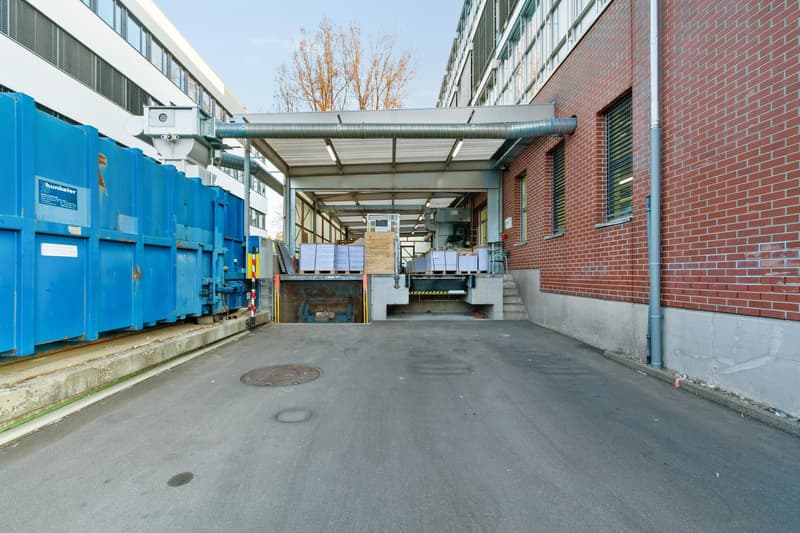 An industrial building with a covered loading bay, surrounded by brick walls and a concrete path, containing blue dumpsters, steel structures, and pallets.