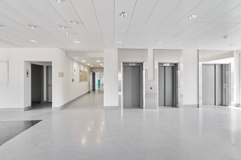 Empty hallway with white walls, light gray flooring, 3 elevators, white ceiling, recessed lighting