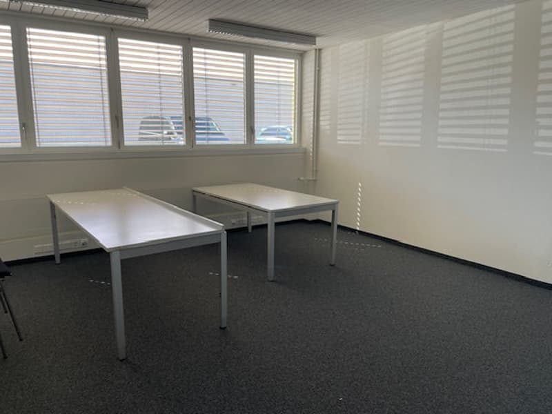 The image shows an office space with two desks, a carpeted floor, and large windows providing natural light. The room appears to be a simple, functional office space.