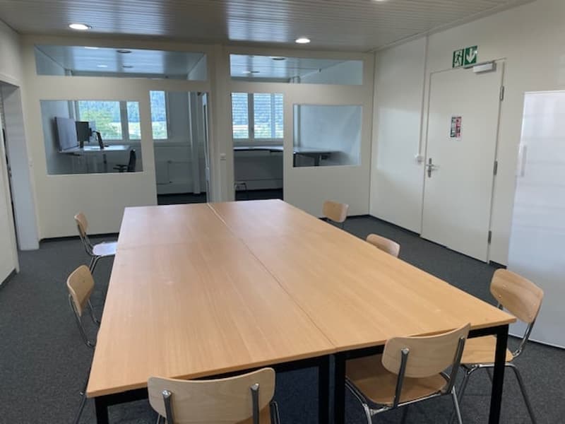 The image shows a modern office space with a large wooden conference table surrounded by chairs. The room has glass walls and doors, allowing natural light to enter. There is a TV or monitor mounted on the wall, and the space appears to be well-equipped f