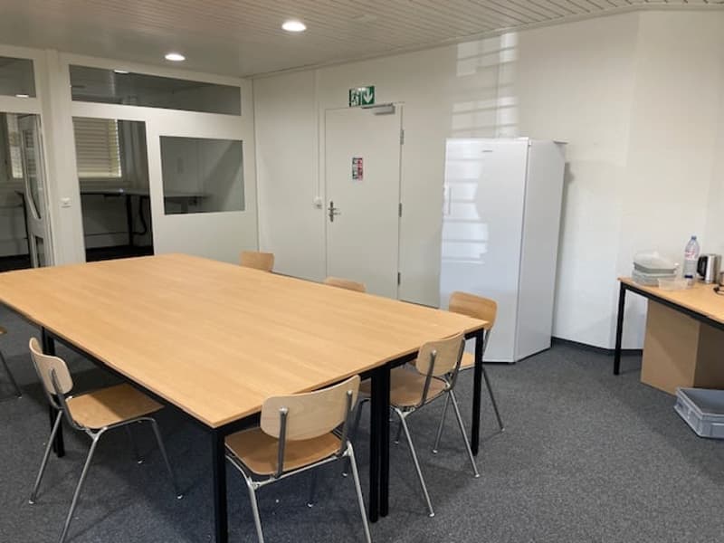 Large wooden table with chairs, refrigerator, and exit door in a modern office space with white walls and recessed lighting.