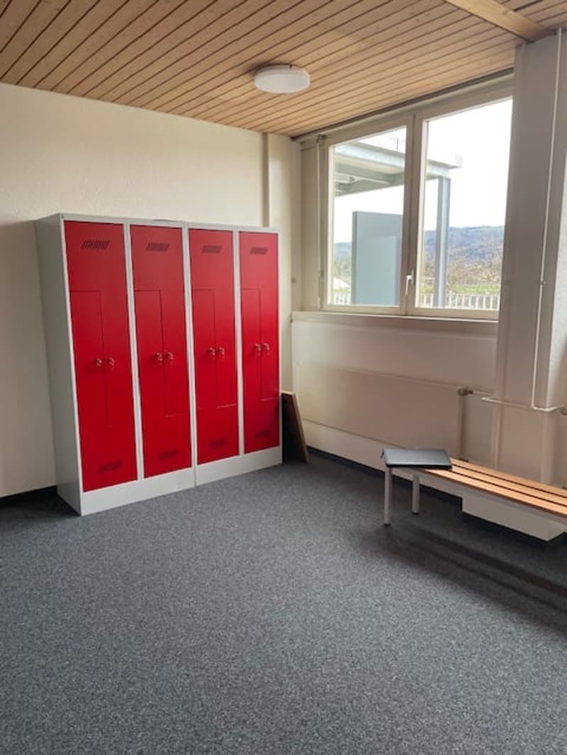 The image shows a locker room with four red lockers, a bench, and a window with a view outside.
