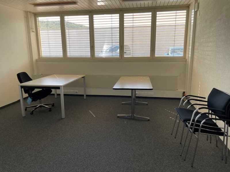 The image shows a small office space with several desks and chairs. The room has large windows that provide natural light, and the walls are painted a neutral color. The floor is covered in a dark carpet. The space appears to be set up for meetings or ind