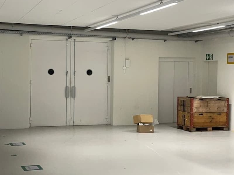 The image shows a large, open warehouse space with white walls, a concrete floor, and several large metal doors. There are some boxes and crates visible in the room, indicating it is being used for storage or distribution purposes.
