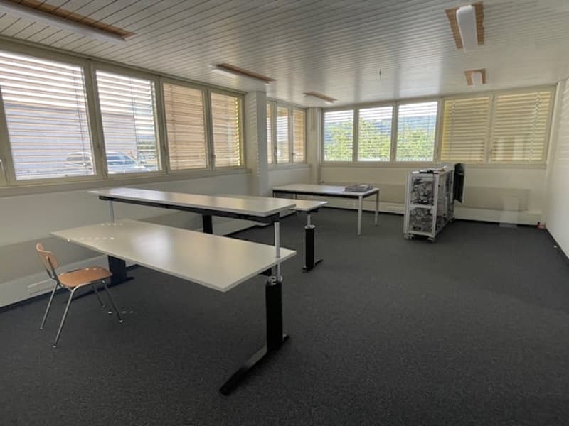 Large open office space with adjustable desks, windows along the walls, and a storage unit in the corner