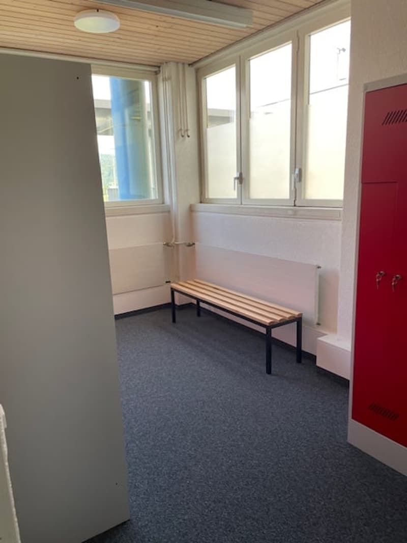 The image shows a hallway with a wooden ceiling, white walls, and a bench along one side. There are large windows that provide natural light, and a red locker or storage unit can be seen in the corner.