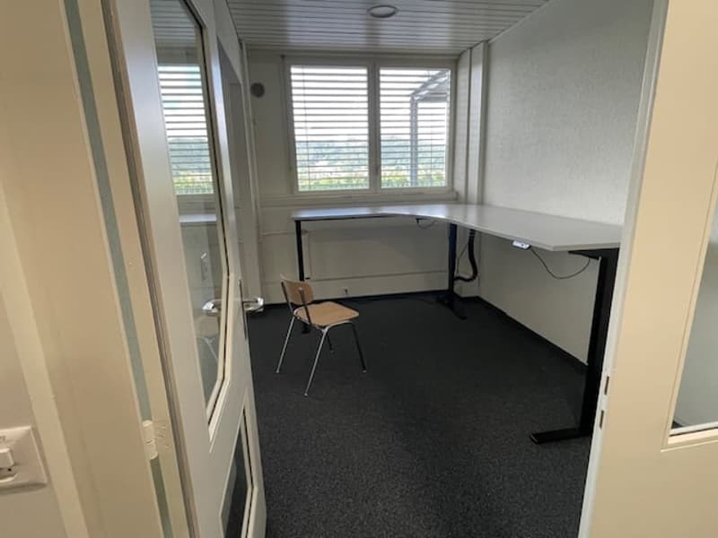 The image shows a small office space with a desk, chair, and large windows providing natural light. The room has a simple, minimalist design with gray walls and dark carpeting.