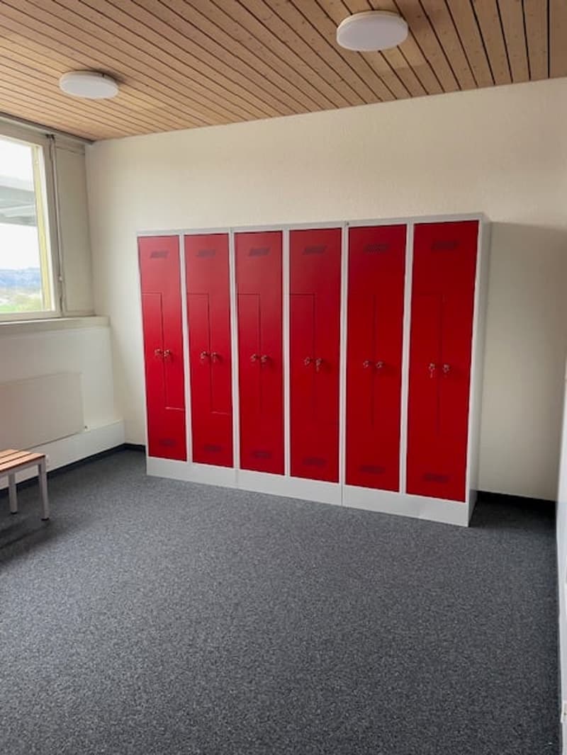 The image shows a room with a row of six red lockers against a white wall. The ceiling has a wooden paneled design with recessed lighting. The floor is covered in gray carpeting.