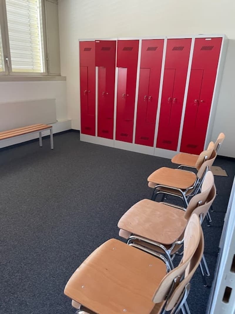 The image shows a locker room with several red lockers lining the wall. There is a bench in front of the lockers and some chairs nearby, suggesting this is a space for changing or storing personal items.