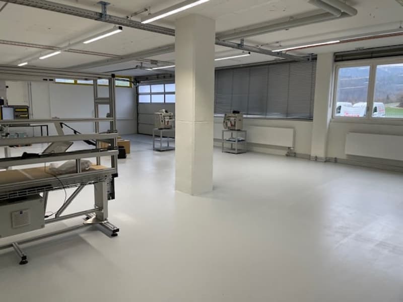 Large open industrial space with concrete floors, metal shelving units, and large windows providing natural light. The space appears to be used for storage or manufacturing purposes.
