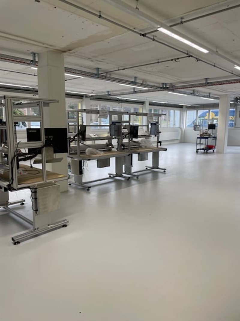Large open office space with multiple workstations, computers, and adjustable desks. The space has high ceilings, bright lighting, and a raised floor.