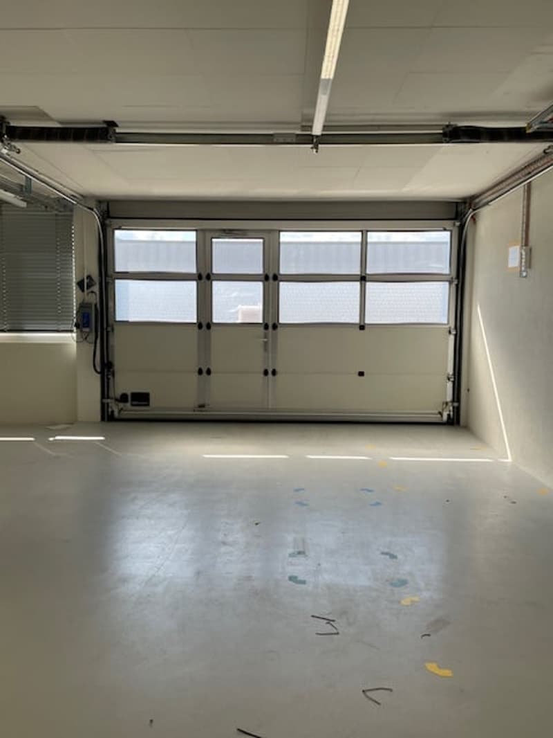 The image shows the interior of a garage with a large garage door, concrete floor, and overhead lighting fixtures.