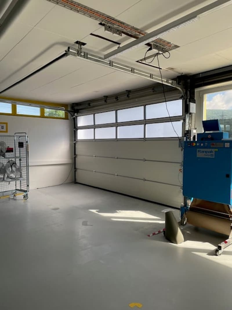 The image shows the interior of a garage. The garage has a large garage door with windows, a concrete floor, and various storage and equipment items visible.