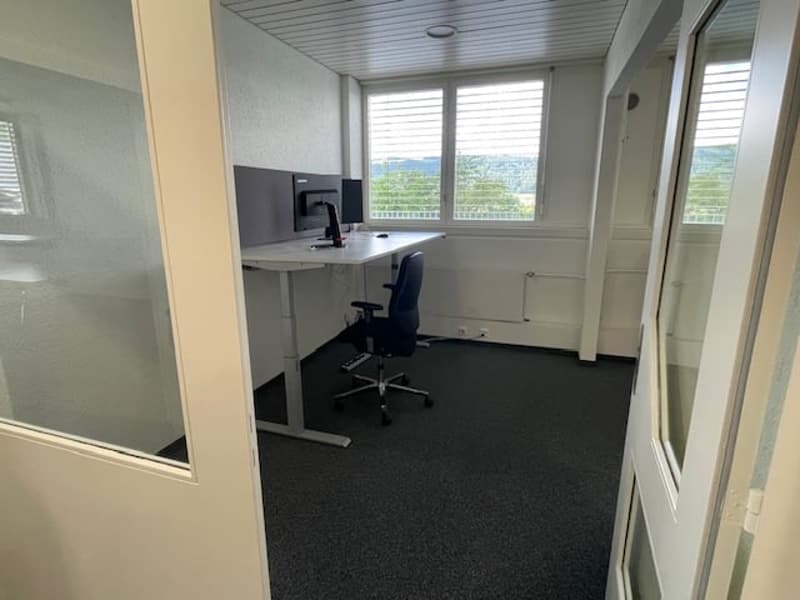 The image shows a small office space with a desk, computer, and office chair. The room has large windows that provide a view of the outdoors. The floor is covered in dark carpeting, and the walls are painted a light color.