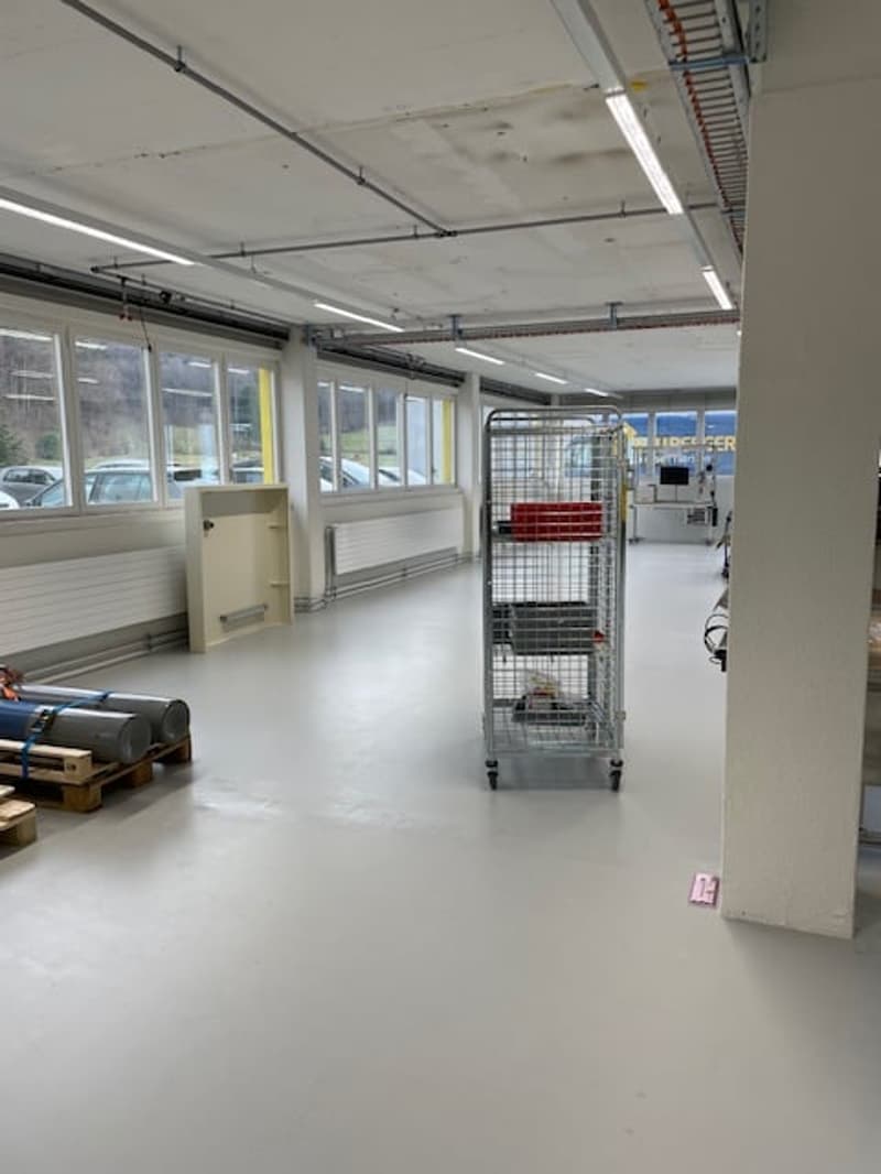 The image shows an open office space with a concrete ceiling, fluorescent lighting, and large windows. There is a metal storage rack and some other equipment visible in the room.