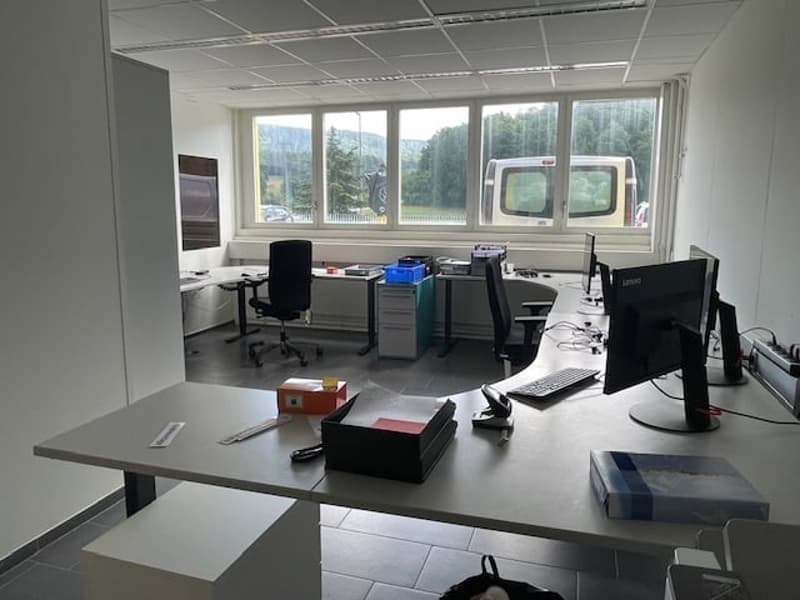 The image shows an office space with large windows providing a view of the outdoors. The room has a desk, computer monitors, and other office equipment and supplies.