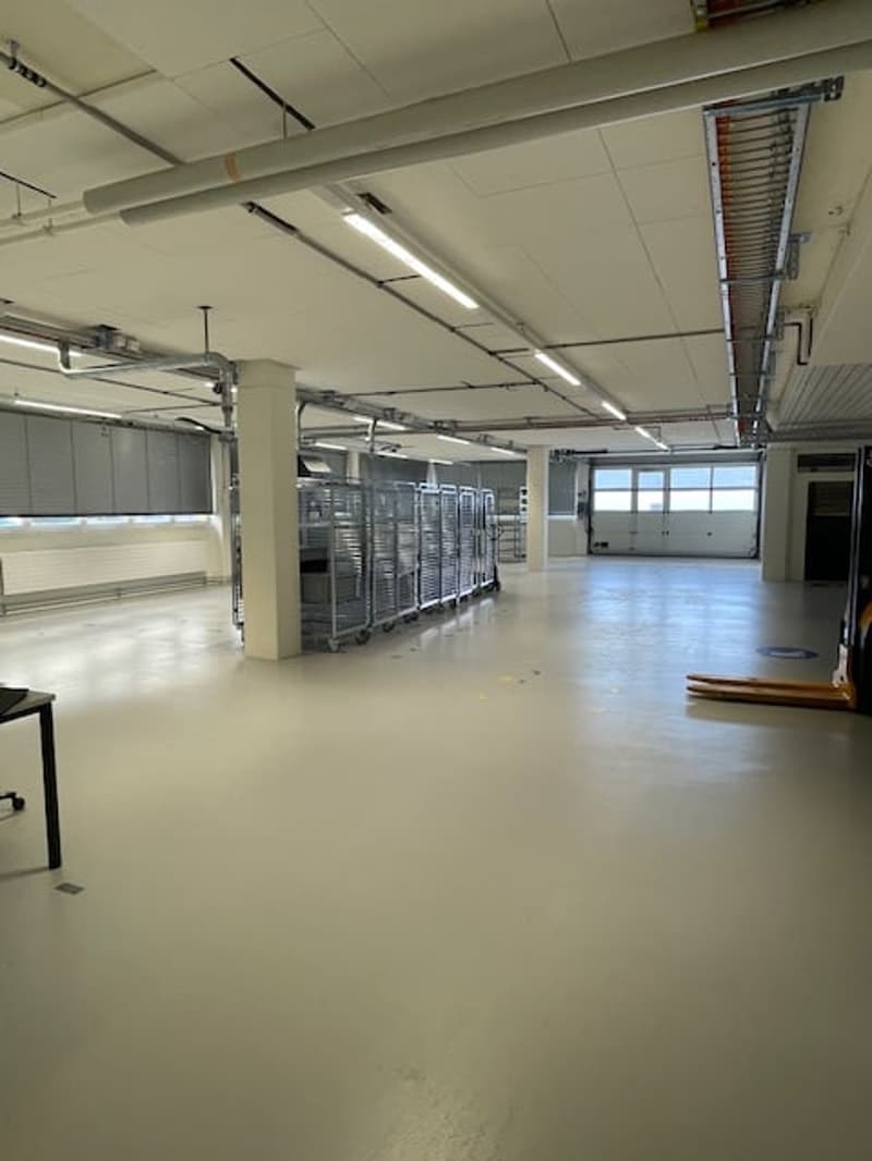 Large open space with concrete floor, metal shelving units, and overhead lighting. The space appears to be a warehouse or storage facility.