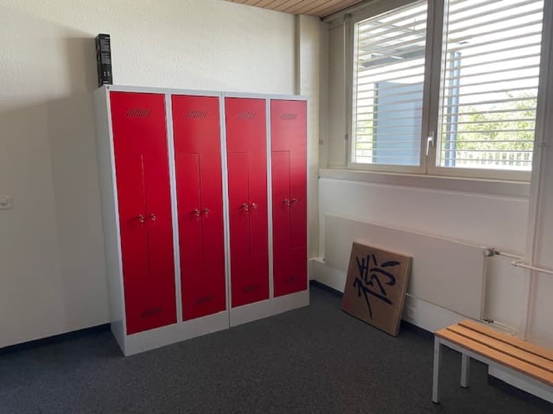 The image shows a room with four red lockers or storage units against the wall. There is a bench or seating area in front of the lockers, and a window with blinds. The floor appears to be carpeted.