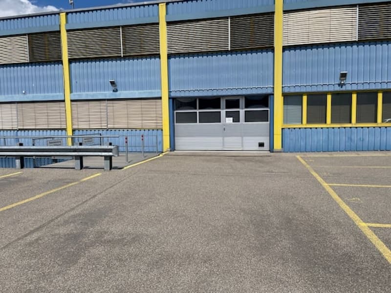 Large industrial building with blue and yellow metal siding, multiple garage doors, and a paved parking area in the foreground.