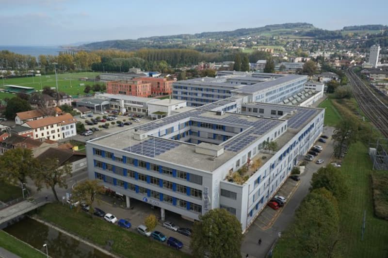 Large multi-story office or commercial building complex with a modern, rectangular design and blue-tinted windows. The building is surrounded by parking lots and green spaces, with a view of a hilly, residential area in the background.