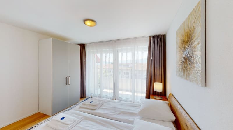 Modern bedroom with 2 single beds, white wardrobe, ceiling lamp, nightstand, curtains, decorative lamp, large glass windows with terrace view, wooden floor.