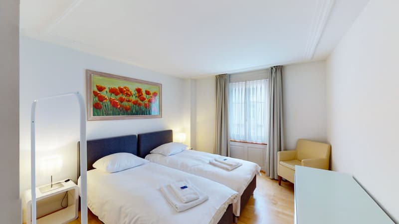 Two beds, white towels, two nightstands with lamps, beige armchair, framed picture, large window