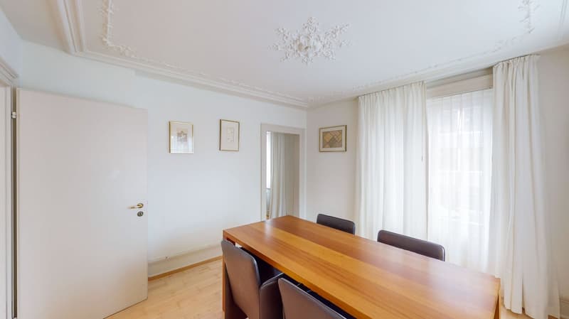 An empty dining room with a wooden table, chairs, and white walls, along with an arched doorway leading to another room. Two windows on the wall, adorned with white curtains. Three picture frames are mounted on the wall above the table.