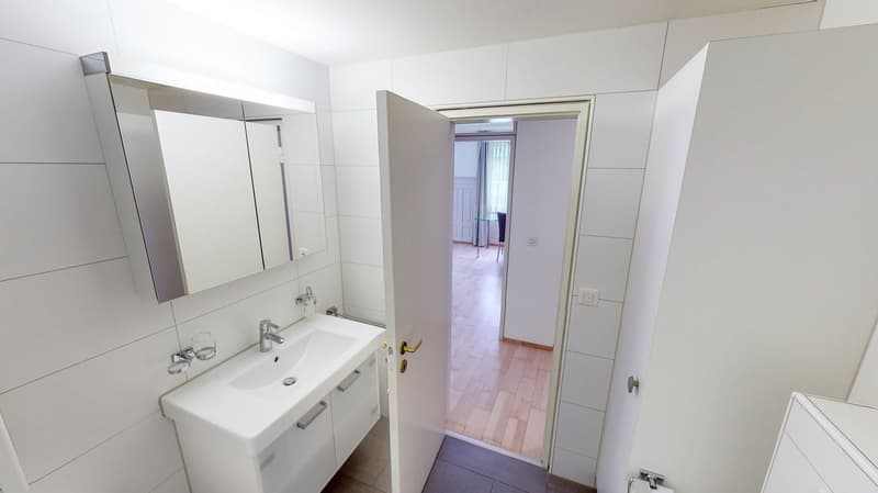 Modern bathroom with white tiled walls, mirror with silver frame, white sink, glass holder, wooden floor, white door