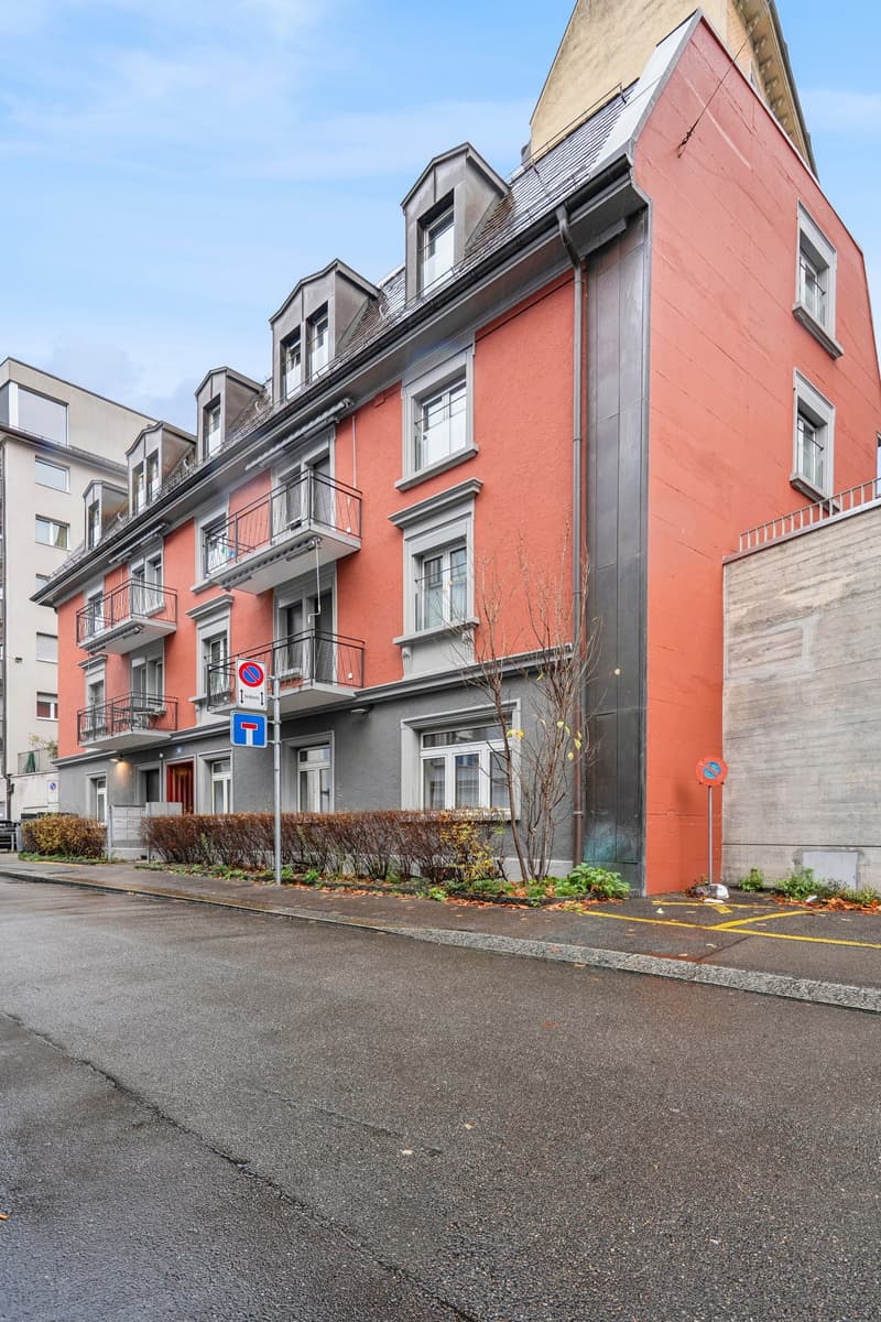 multi-story building with red brick facade, multiple windows, balconies, and metal piping, several windows on the side facing the street, a grassy area in front with some small plants, an entrance with a door and some stairs leading up to it, other buildi