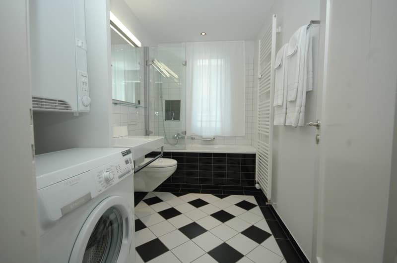 Modern white washer and dryer, white tiled walls, shower and bathtub, toilet, white towels, white radiator