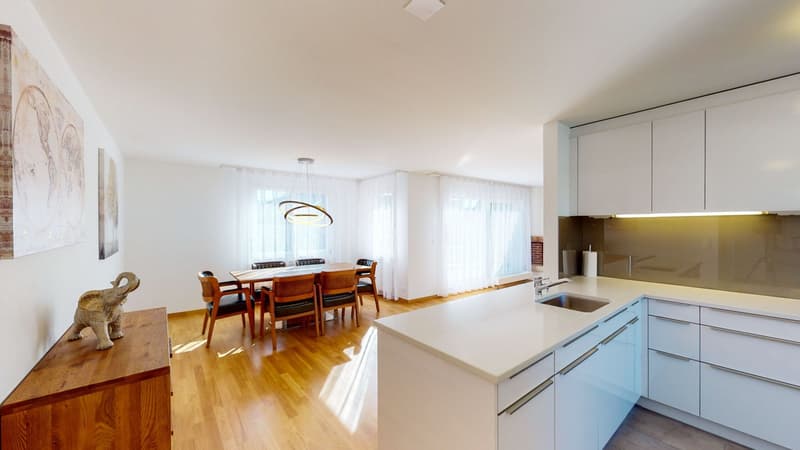 Open kitchen with wooden floor, white countertops, under-cabinet lighting, stainless steel sink, built-in dishwasher, dining table with chairs, decorative items, large windows with white curtains.