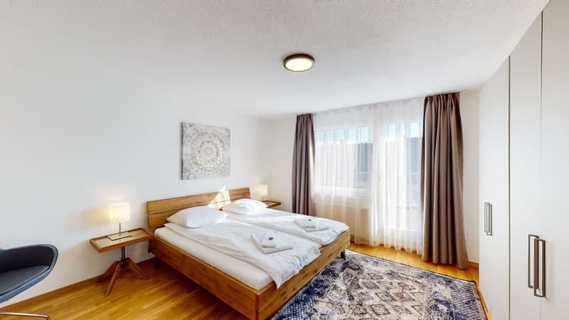 A bedroom with a double bed, nightstands with lamps, a chair, a wooden cabinet, a rug, and a window with curtains.
